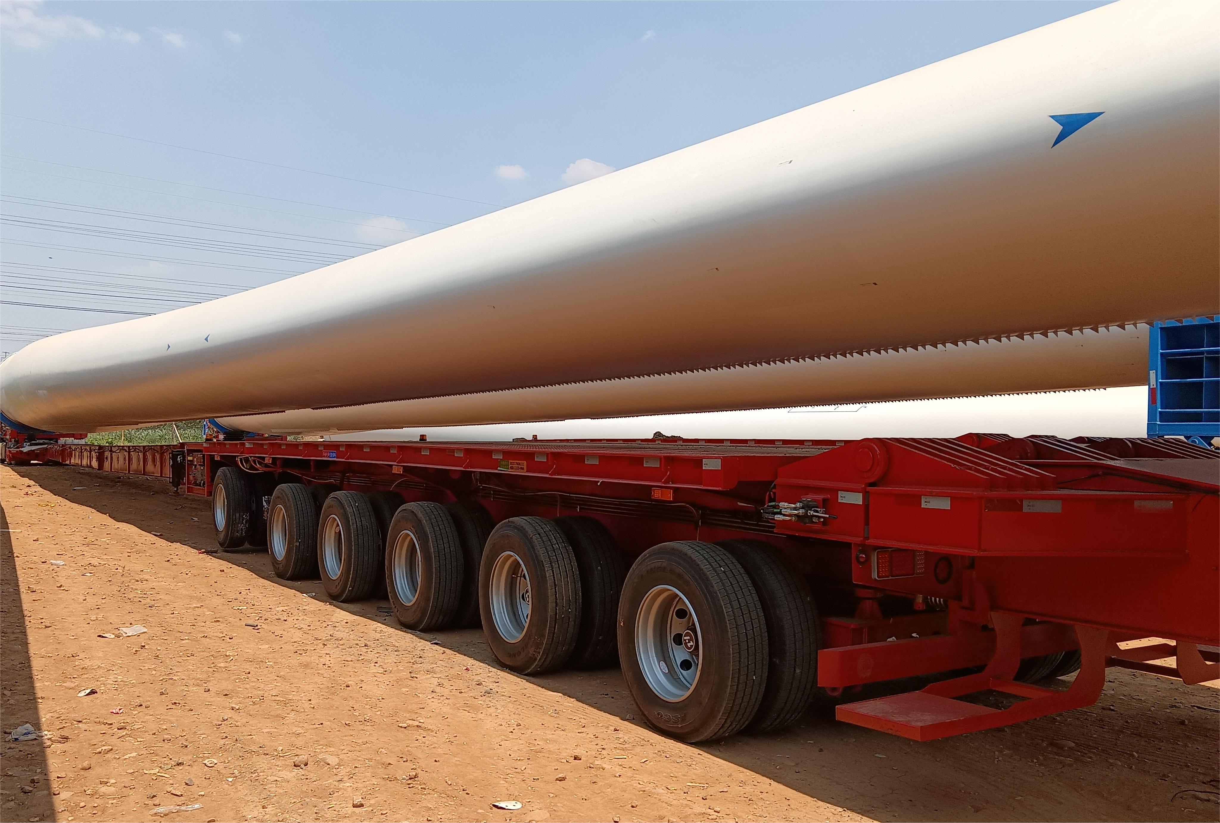A rear-side view photograph capturing a HUAYUE wind blade transporter, loaded with a massive white wind turbine blade. This client feedback photo highlights the stable rear axle design during a challenging turn, verifying maneuverability despite extreme length
