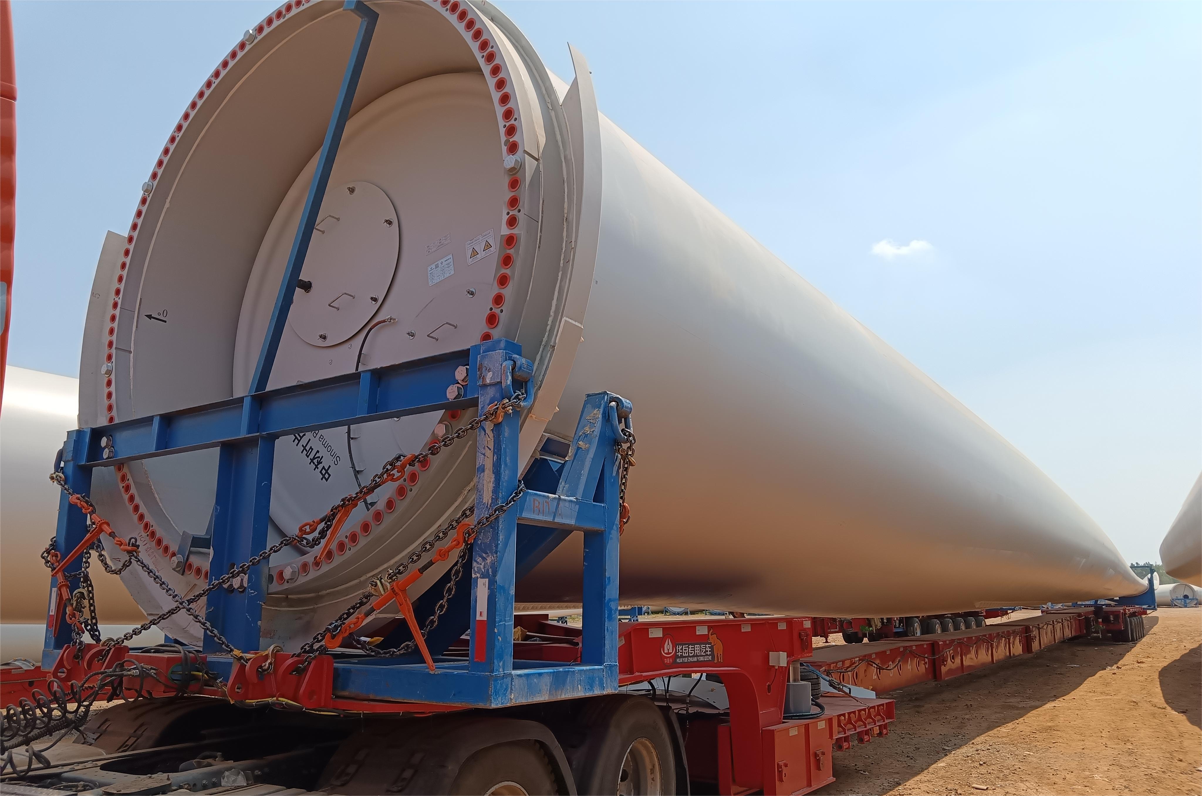 A side view photograph of a red prime mover towing a HUAYUE trailer loaded with a massive white wind turbine blade on a public road. This is a client feedback photo demonstrating successful loading and stable transport