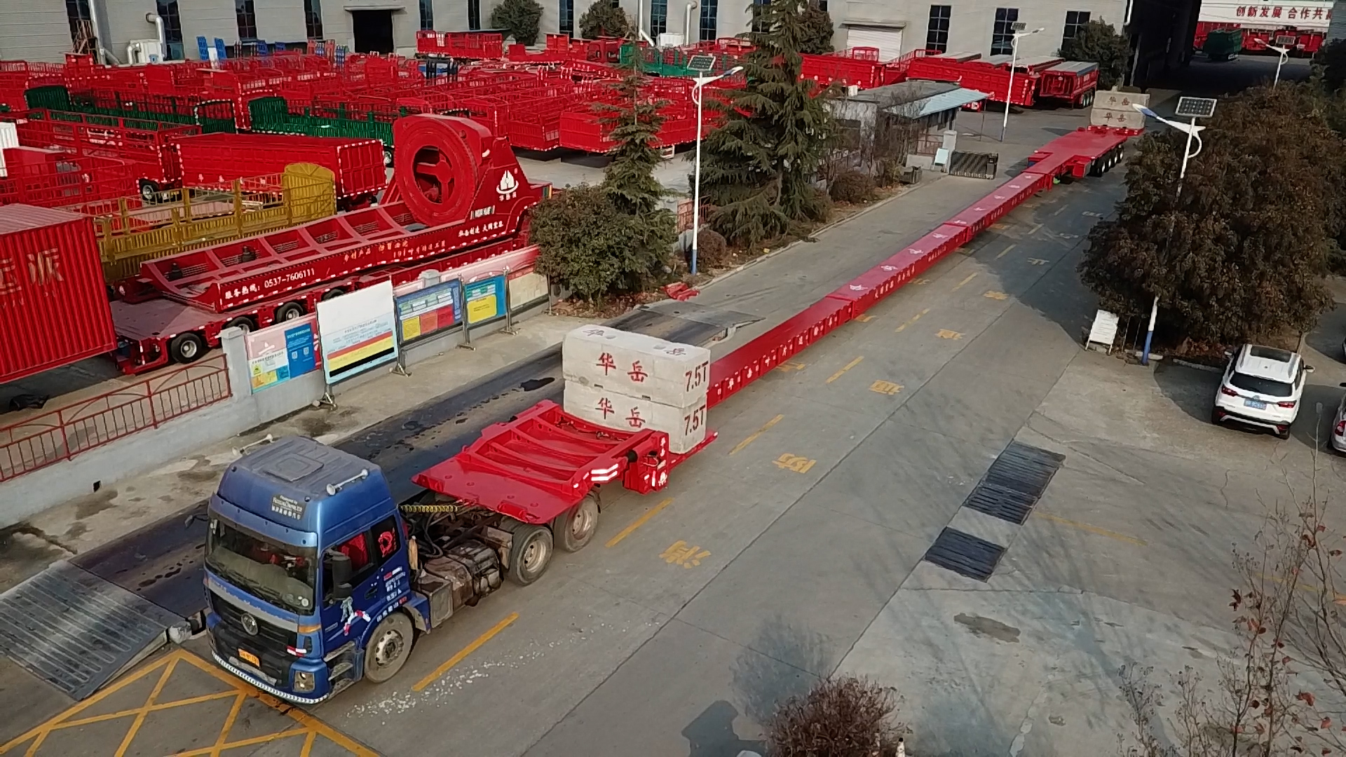 An aerial photograph looking down at the HUAYUE factory yard, capturing the immense scale of an empty, red expandable wind blade transporter trailer, showcasing its extreme extended length capabilities for hauling super-long cargo.