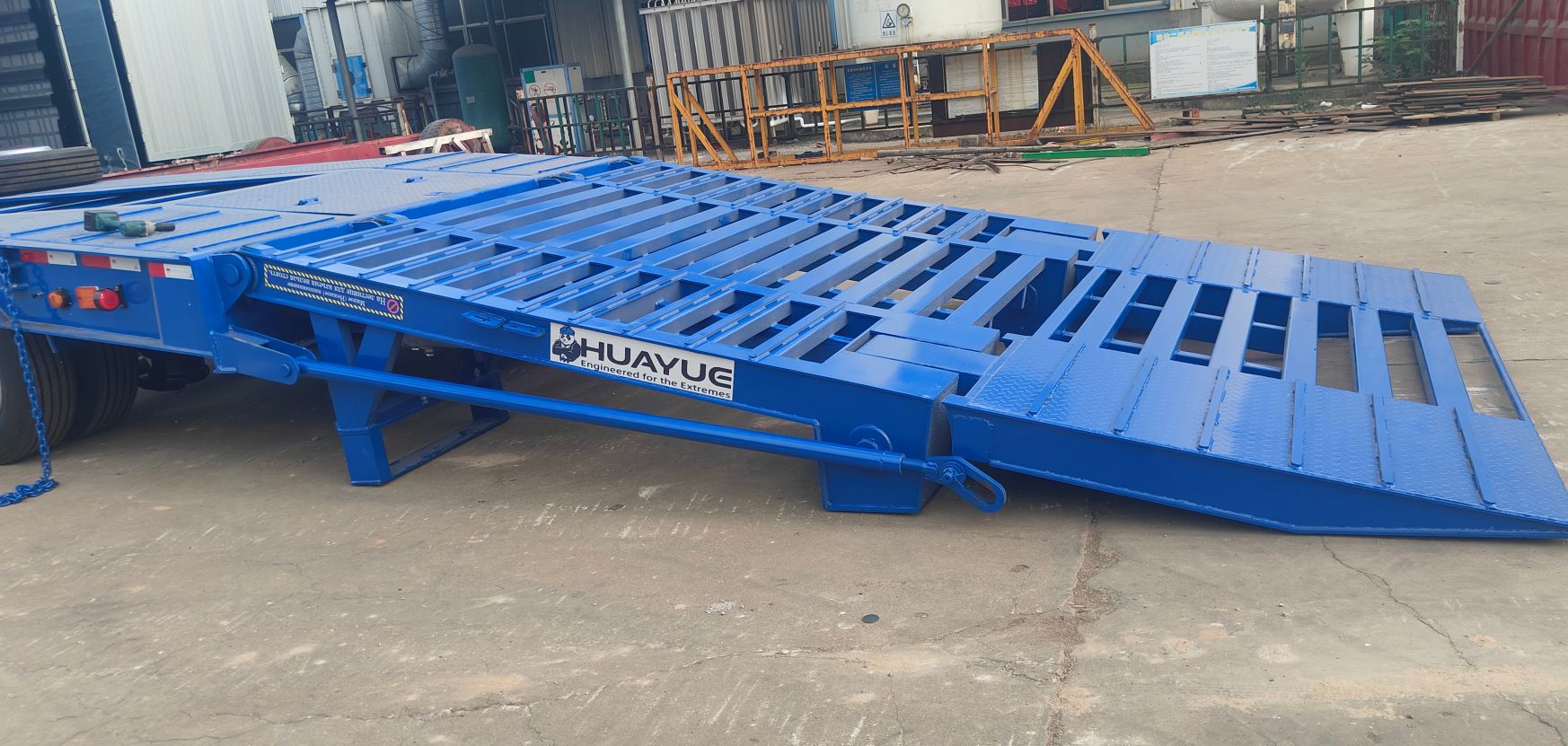 Side close-up of the HUAYUE lowbed trailer's double-folding hydraulic ramps. Clearly displaying the heavy-duty hydraulic cylinder support structure and the extremely low climbing angle formed when fully deployed, effectively preventing the seesaw effect during excavator loading.